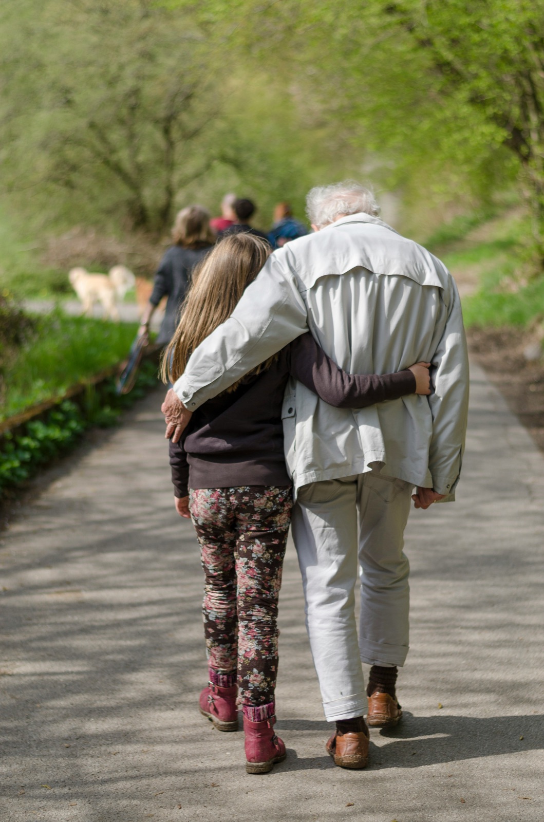 Ein älterer Mann und ein Kind gehen Arm in Arm einen Weg entlang, von hinten gesehen.