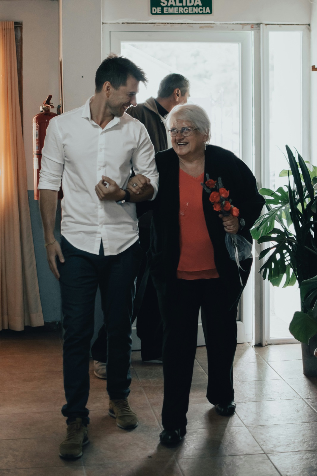 Ein junger Mann und eine ältere Frau mit Blumen in der Hand gehen untergehakt nebeneinander.